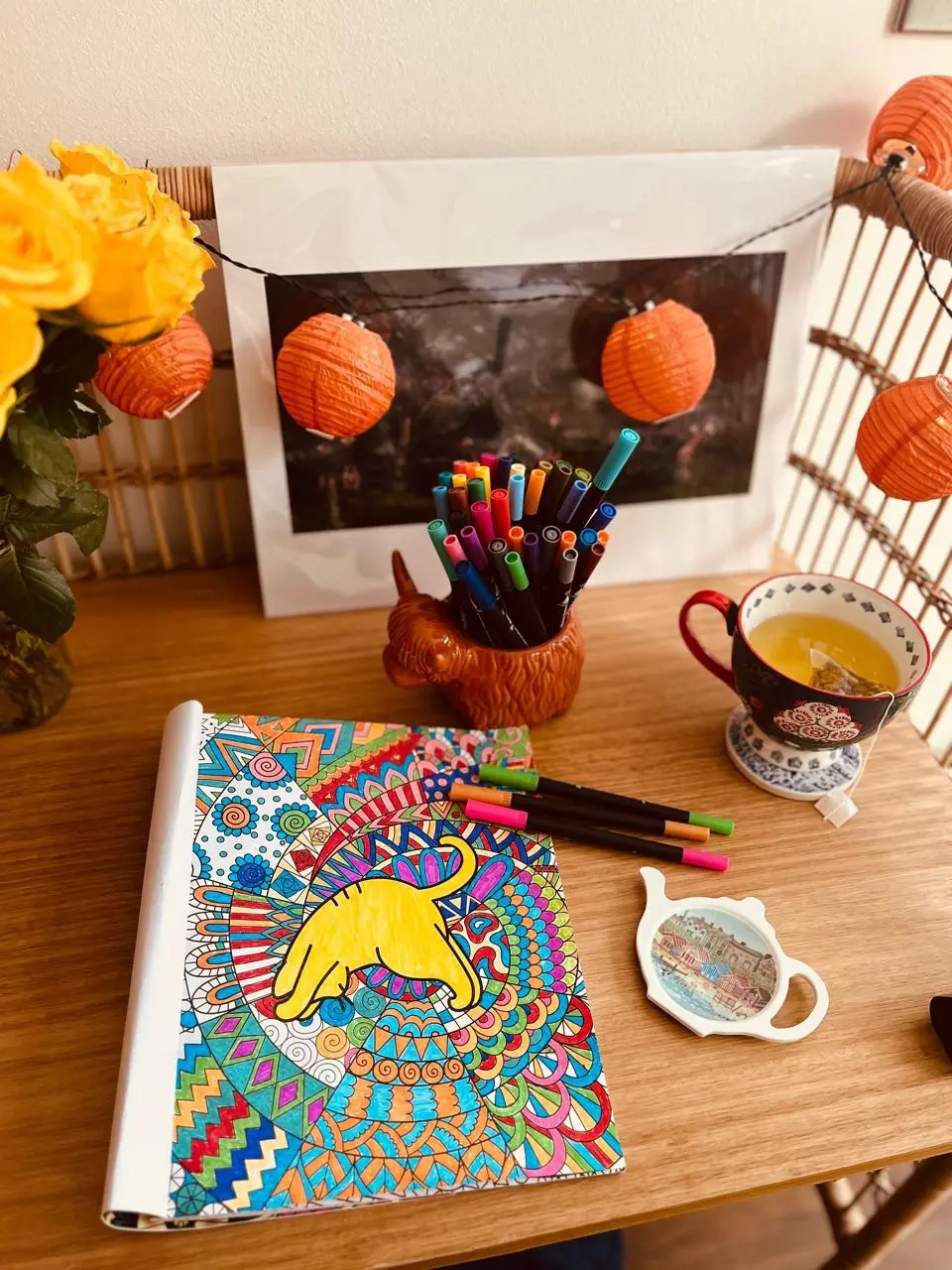 A desk with a part-coloured mandala of a lion, a jar of felt pens, a cup of tea and a vase of yellow roses